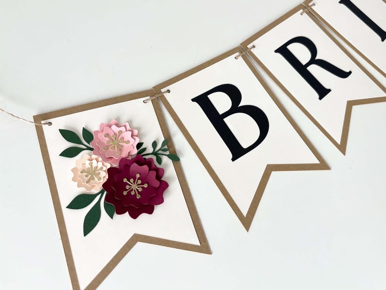 Op de afbeelding: Een decoratieve slinger bestaande uit witte kartonnen vlaggetjes met bruine randen aan een touwtje. E&eacute;n vlaggetje is versierd met driedimensionale papieren bloemen in roze, bordeaux en groen. Andere vlaggetjes bevatten grote zwarte hoofdletters.