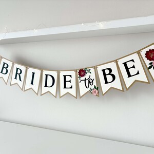 Op de afbeelding: Een decoratieve slinger met witte wimpels met gouden randen en zwarte letters die BRIDE TO BE spellen. De slinger is versierd met driedimensionale papieren bloemen in tinten donkerrood, lichtroze en groen blad.