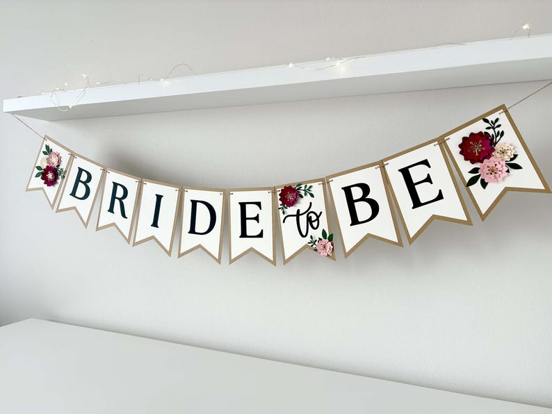 Op de afbeelding: Een decoratieve slinger met witte wimpels met gouden randen en zwarte letters die BRIDE TO BE spellen. De slinger is versierd met driedimensionale papieren bloemen in tinten donkerrood, lichtroze en groen blad.