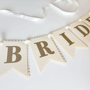 May include: A cream-colored banner with gold lettering spelling "BRIDE" and a ring icon. The banner is made of individual pennant-shaped flags with scalloped edges, strung together with a white ribbon. Ideal for a bridal shower or wedding.