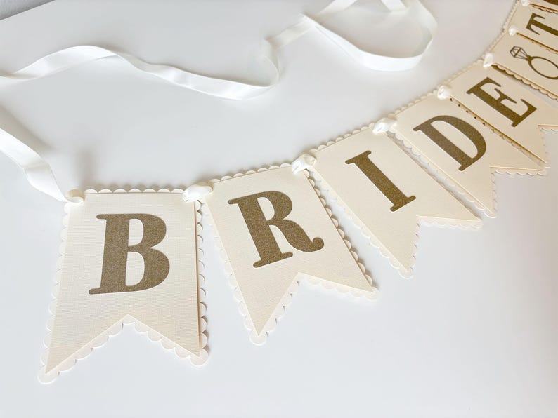 May include: A cream-colored banner with gold lettering spelling "BRIDE" and a ring icon. The banner is made of individual pennant-shaped flags with scalloped edges, strung together with a white ribbon. Ideal for a bridal shower or wedding.