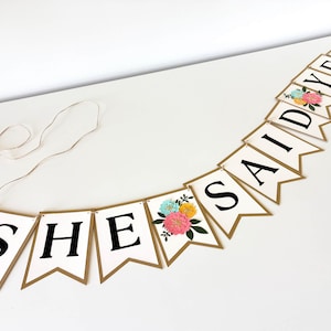 Op de afbeelding: Een decoratieve slinger met witte wimpels met gouden randen, elk voorzien van een zwarte letter die samen SHE SAID YES vormen. Twee wimpels zijn voorzien van een bloemenillustratie met roze, gele en blauwe bloemen en groene bladeren.