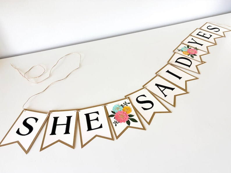 Op de afbeelding: Een decoratieve slinger met witte wimpels met gouden randen, elk voorzien van een zwarte letter die samen SHE SAID YES vormen. Twee wimpels zijn voorzien van een bloemenillustratie met roze, gele en blauwe bloemen en groene bladeren.
