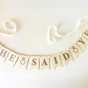 May include: A banner with the words "SHE SAID YES" printed in brown on cream-colored pennants. Each pennant is strung together with a white ribbon and small white beads. A diamond ring graphic is also included.