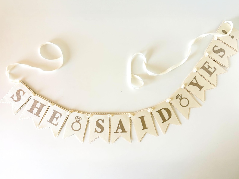 May include: A banner with the words "SHE SAID YES" printed in brown on cream-colored pennants. Each pennant is strung together with a white ribbon and small white beads. A diamond ring graphic is also included.