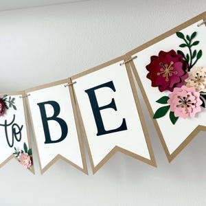 Op de afbeelding: Een decoratieve slinger met witte wimpels met bruine randen aan een touwtje. De wimpels tonen de zwarte tekst E, to, B, E, en een laatste wimpel is versierd met gelaagde papieren bloemen in donkerrood, lichtroze en cr&egrave;me met groene bladeren.