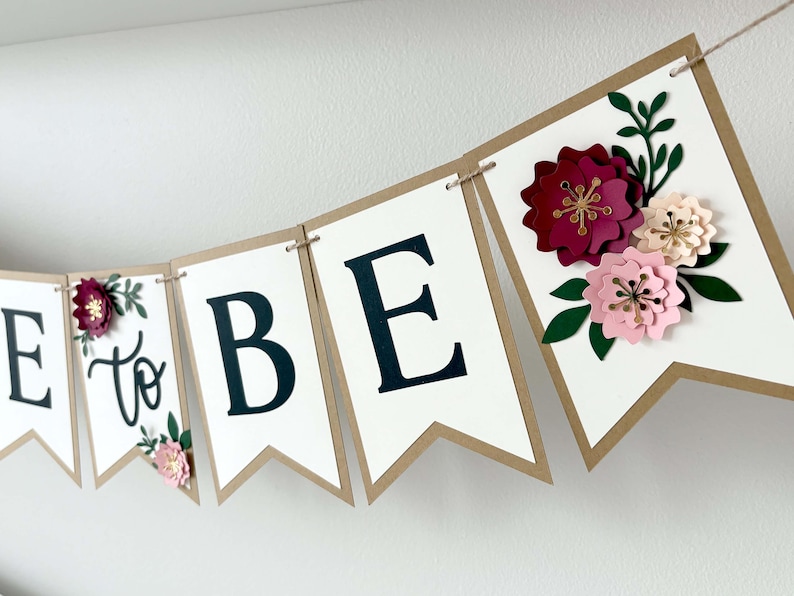 Op de afbeelding: Een decoratieve slinger met witte wimpels met bruine randen aan een touwtje. De wimpels tonen de zwarte tekst E, to, B, E, en een laatste wimpel is versierd met gelaagde papieren bloemen in donkerrood, lichtroze en cr&egrave;me met groene bladeren.