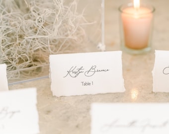 May include: A wedding table setting with place cards. Each white card has a torn edge, with a name in cursive and a table number. A lit candle in a glass holder and decorative elements are in the background. The setting is elegant.