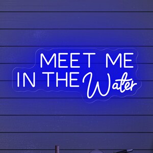 May include: A blue neon sign with the words "MEET ME IN THE Water" in a cursive font. The sign is mounted on a blue wooden wall. Decorative nautical items are visible below the sign, including a ship's wheel and a bottle.