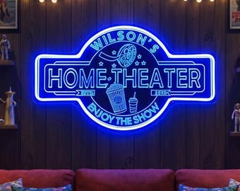 May include: A home cinema setup with a red sofa, vintage film posters, and a neon sign reading "Wilson's Home-Theater." String lights and decorative cushions enhance the cosy atmosphere. Popcorn and drinks are on side tables.