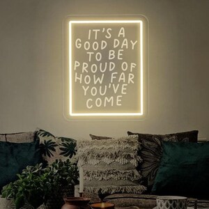 May include: A white neon sign with a black frame that reads "It's a good day to be proud of how far you've come". The sign is hanging on a wall above a couch with pillows.
