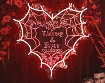 Puede incluir: Una pareja posa en un sofá de terciopelo rojo en un entorno de bosque oscuro. Un letrero de neón en forma de corazón con un diseño de telaraña dice "Lindsey & Ryan 03.12.2025".