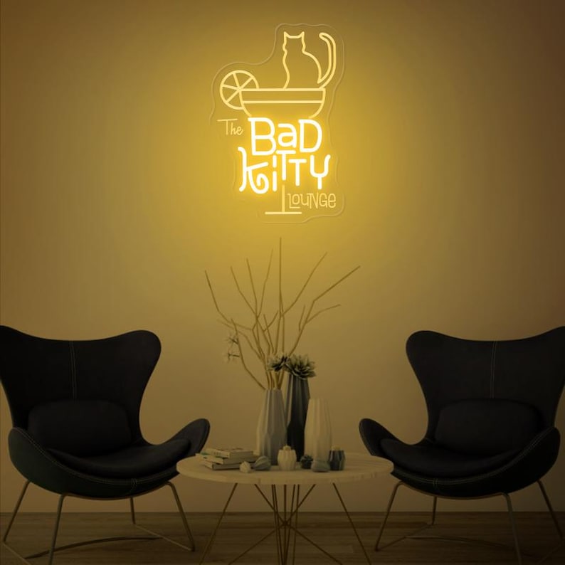 Peut inclure: Enseigne au n&eacute;on jaune repr&eacute;sentant un chat dans un verre &agrave; cocktail, avec l'inscription "The Bad Kitty Lounge". L'enseigne est fix&eacute;e au mur au-dessus d'une petite table et de deux fauteuils noirs.