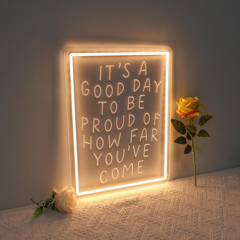 May include: A white neon sign with the text "It's a good day to be proud of how far you've come". The sign is lit up and is sitting on a white surface with a yellow flower next to it.
