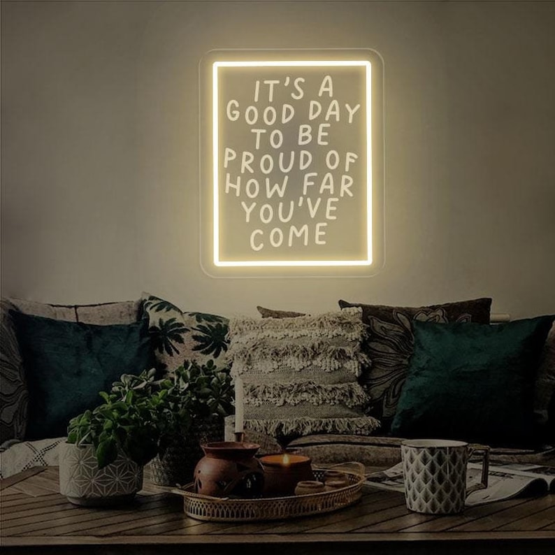May include: A white neon sign with a black frame that reads "It's a good day to be proud of how far you've come". The sign is hanging on a wall above a couch with pillows.
