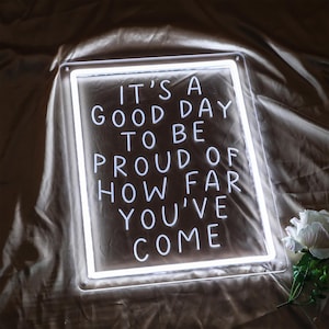 May include: White neon sign with the text "It's a good day to be proud of how far you've come".