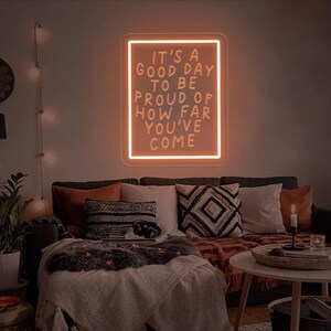 May include: A neon sign with the text "It's a good day to be proud of how far you've come" in a white frame against a dark wall. The sign is lit up in a warm orange color.