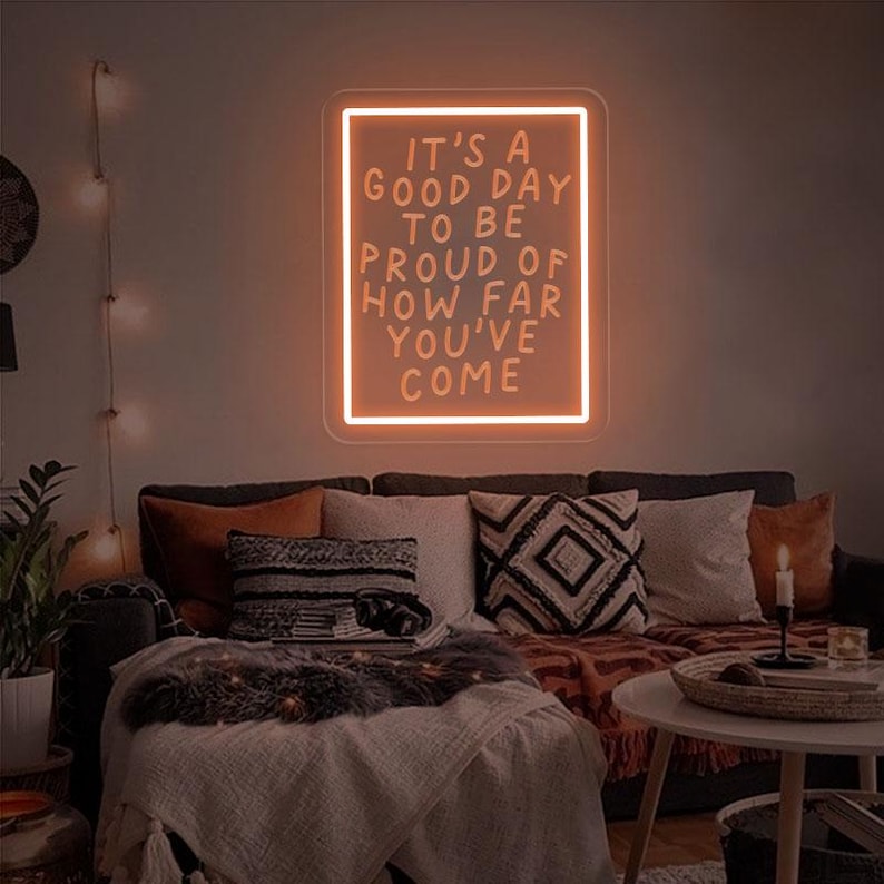 May include: A neon sign with the text "It's a good day to be proud of how far you've come" in a white frame against a dark wall. The sign is lit up in a warm orange color.