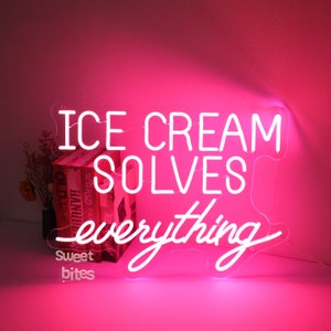 May include: White neon sign with pink lighting that reads "ICE CREAM SOLVES everything" with the word "everything" in cursive. The sign is mounted on a pink wall and there are books below the sign.