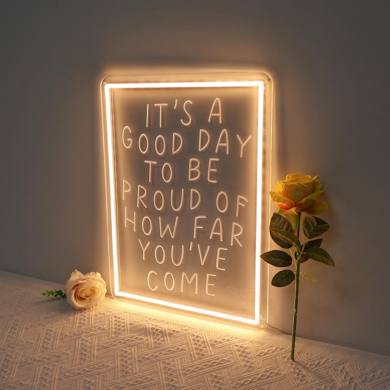 May include: A white neon sign with the text "It's a good day to be proud of how far you've come". The sign is illuminated and has a white frame. The sign is sitting on a white surface with a yellow rose in the foreground.