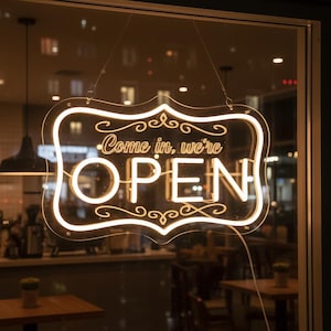 May include: A neon sign with the words "Come in, we're OPEN" in a decorative font. The sign is outlined in a warm, glowing light and hangs in a window, inviting customers. The background shows a glimpse of a cafe interior.