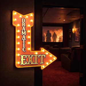 May include: An illuminated, arrow-shaped sign with the words "DRAMATIC EXIT" in a retro font. The sign is orange with yellow trim and lights, mounted on a dark wall. The arrow points to the right, suggesting an exit.