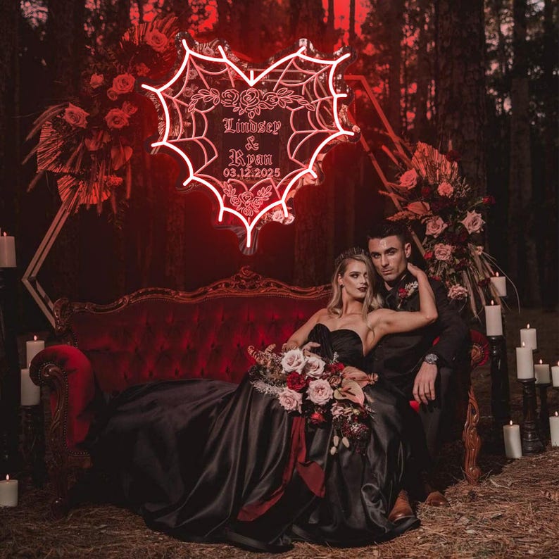 Peut inclure: Un couple pose sur un canap&eacute; en velours rouge dans un d&eacute;cor de for&ecirc;t sombre. Un n&eacute;on en forme de c&oelig;ur avec un motif de toile d'araign&eacute;e indique "Lindsey & Ryan 03.12.2025".