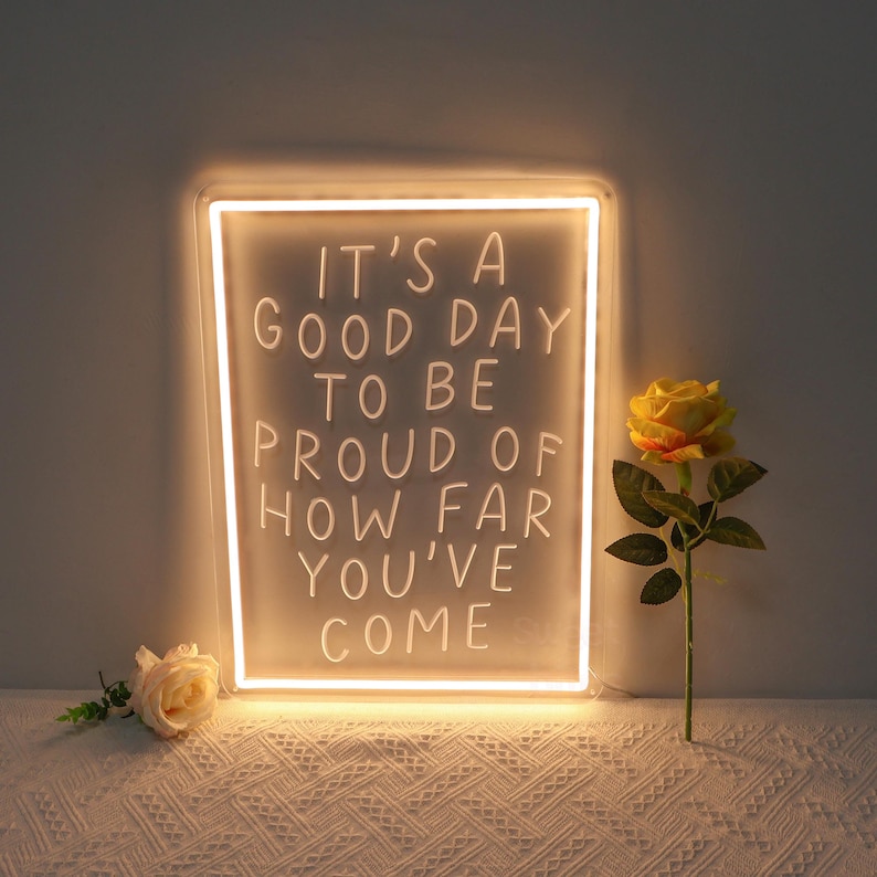 May include: A white neon sign with the text "It's a good day to be proud of how far you've come". The sign is lit up and is sitting on a white surface with a yellow rose in the background.