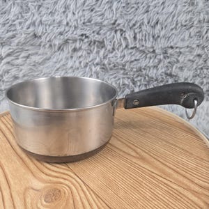 May include: A stainless steel saucepan with a dark, textured handle. The handle is attached to the side of the pan with two visible metal screws and features a small, circular metal loop at the end for hanging the cookware in a kitchen.
