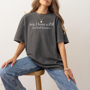 May include: Dark gray t-shirt with the text "yes, I have a rbf (red bull fixation)" in white. The shirt is a relaxed fit and is paired with blue jeans. The model is wearing gold necklaces and bracelets.
