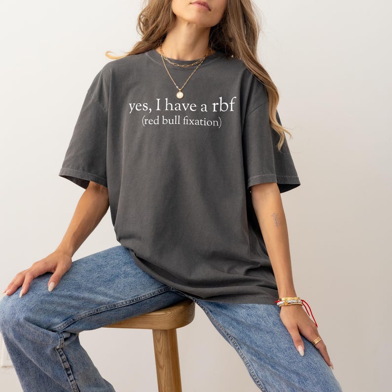 May include: Dark gray t-shirt with the text "yes, I have a rbf (red bull fixation)" in white. The shirt is a relaxed fit and is paired with blue jeans. The model is wearing gold necklaces and bracelets.