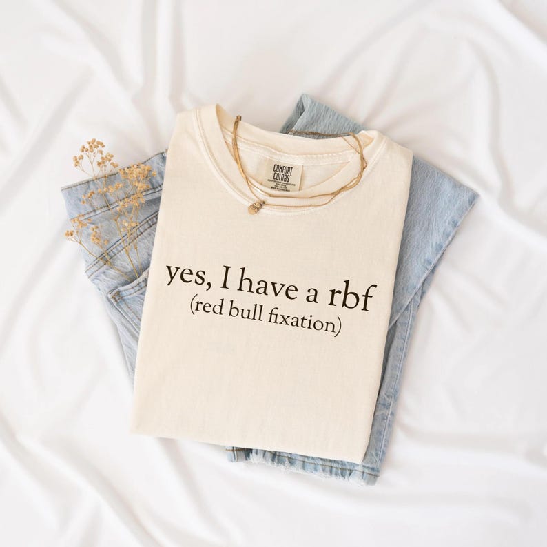 May include: Cream-colored t-shirt with the text "yes, I have a rbf (red bull fixation)". The shirt is folded and placed on top of a pair of light blue jeans. A gold necklace and dried flowers are also visible.