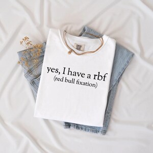 May include: White t-shirt with the text "yes, I have a rbf (red bull fixation)". The shirt is folded on top of a pair of blue jeans. A gold necklace and a sprig of dried flowers are also visible.