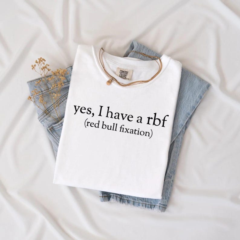 May include: White t-shirt with the text "yes, I have a rbf (red bull fixation)". The shirt is folded on top of a pair of blue jeans. A gold necklace and a sprig of dried flowers are also visible.