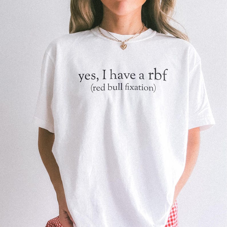 May include: White t-shirt with the text "yes, I have a rbf (red bull fixation)" in black. The shirt has short sleeves and a crew neck. The text is centered on the front of the shirt.