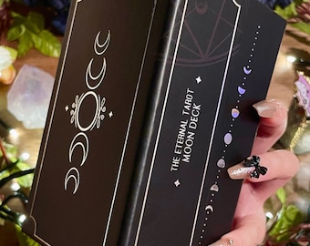 Op de afbeelding: Een zwarte tarotkaartdoos met gouden randen en maanfase-ontwerpen. De tekst "THE ETERNAL TAROT MOON DECK" staat op de zijkant. De doos wordt vastgehouden in een hand met versierde nagels, met een bloemenachtergrond.