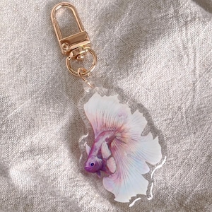 May include: A clear acrylic keychain featuring a colorful betta fish design. The fish has a white, pink, and purple color scheme. The keychain has a gold-colored clasp.