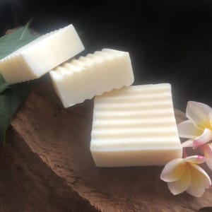 May include: Three bars of white soap with a ribbed texture, arranged on a piece of brown wood with white flowers.