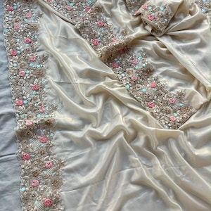 May include: A champagne-coloured satin fabric draped in folds, featuring a wide, intricate border of gold embroidery, small pink fabric roses, and light blue accents. The decorative trim runs along the edges of the smooth, lustrous material.