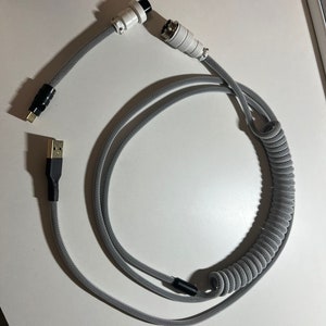 Könnte beinhalten: Ein graues, gewickeltes Kabel mit schwarzen Steckern an beiden Enden. Ein Ende hat einen USB-A-Stecker und das andere Ende eine 3,5-mm-Klinkenbuchse.