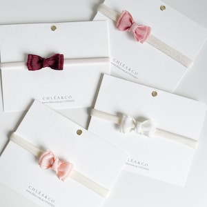 May include: Four elastic headbands, each featuring a small fabric bow in burgundy, dusty rose, white, or light peach. Each headband is attached to a white display card printed with the text CHLÉA & CO. www.chlea.com.au | SYDNEY.