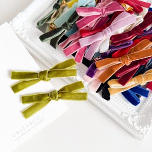 May include: A collection of velvet hair bows in various colours including olive green, pink, red, purple, blue, and tan. Two olive green bows are attached to a white card with the text CHLEA & CO. www.chlea.com.au | SYDNEY printed on it.