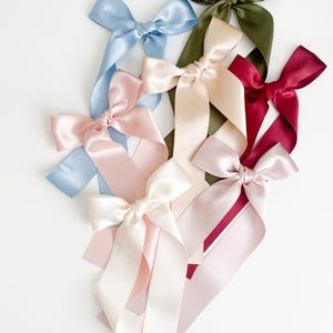 May include: A selection of satin ribbon bows in shades of light blue, pink, cream, olive green, and burgundy. Each bow features long, flowing tails, offering a delicate and elegant appearance. Ideal for gift wrapping or hair accessories, these bows are a charming detail.