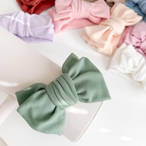May include: A sage green ribbed fabric hair bow featuring a knotted centre design. The bow is displayed on a light-coloured stand, surrounded by a collection of similar hair bows in various pastel colours including pink, lavender, and white.