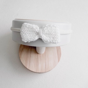 May include: A white knitted bow attached to a thin, matching white elastic headband. The headband is displayed on a smooth, light-coloured wooden stand with a cylindrical base and a flat, rounded top piece that supports the accessory.