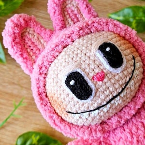 Può includere: Un giocattolo di peluche rosa lavorato a uncinetto con un viso sorridente, occhi neri e un naso rosa. Il giocattolo ha orecchie lunghe e flosce ed è fatto con un filato morbido e peloso.