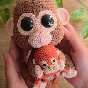 Op de afbeelding: Een handgemaakte gehaakte apenpop in bruin-, beige- en roze tinten, met grote zwarte ogen. De aap houdt een kleinere, oranje aap vast. De pop wordt in de handen van een persoon gehouden.