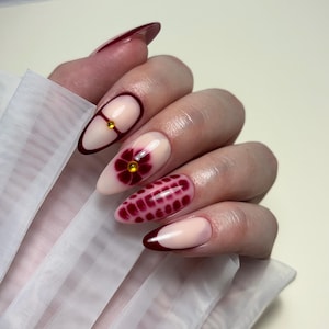 Peut inclure: Une main avec de longs ongles en forme d'amande. Les ongles sont peints avec une variété de motifs, dont une fleur rouge foncé, un motif quadrillé et une manucure française avec une bordure rouge foncé. Certains ongles ont des accents dorés.