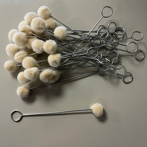 May include: A pile of 20 small, white, fluffy, round, cotton-tipped applicators with silver metal handles. The applicators are arranged in a pile with the handles sticking out. One applicator is laying flat on the table.