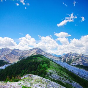 May include: A rocky mountain peak with a green grassy slope and a blue sky with white clouds. The mountain is covered in rocks and vegetation.